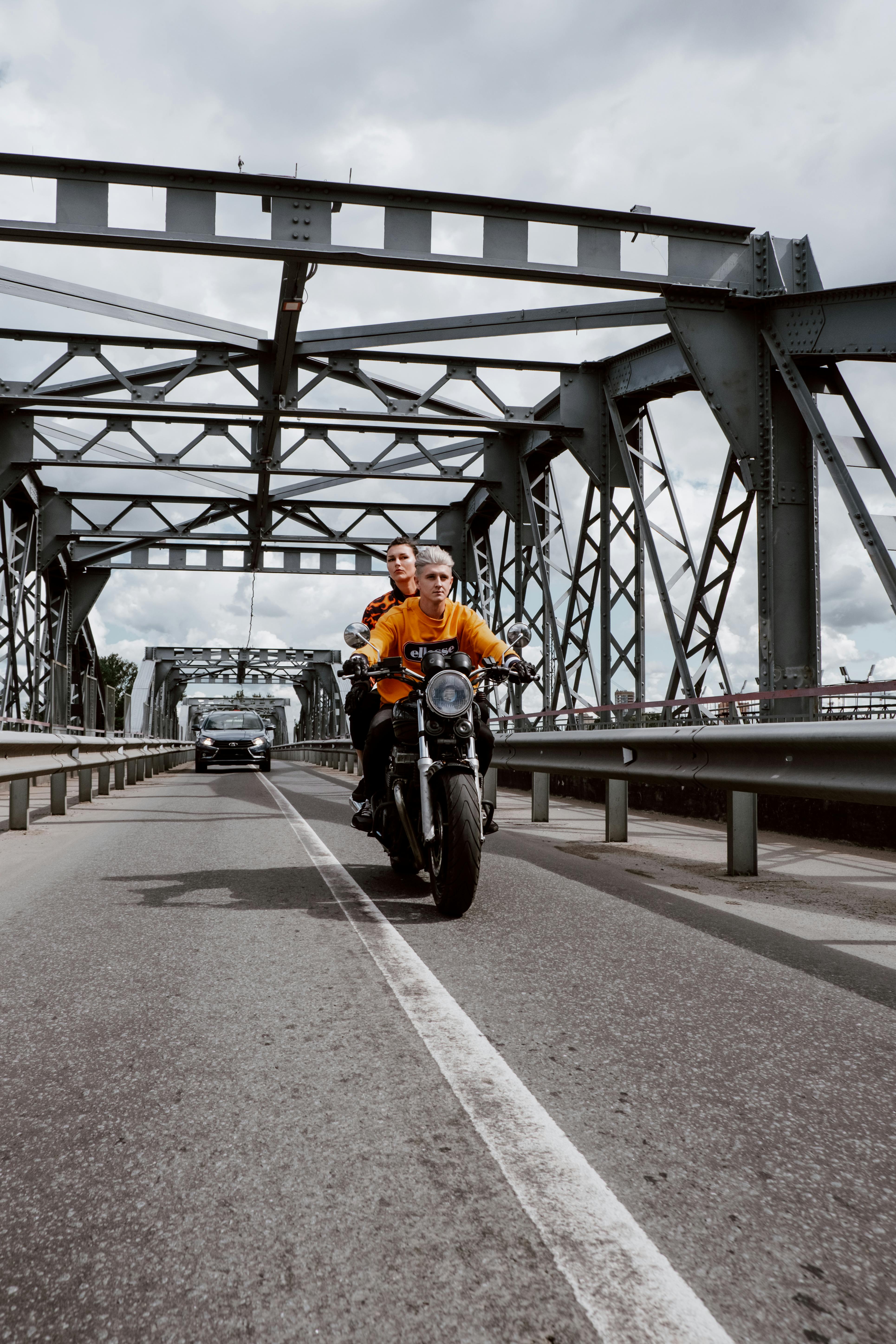A couple riding a motorcycle across a steel bridge with a car following.