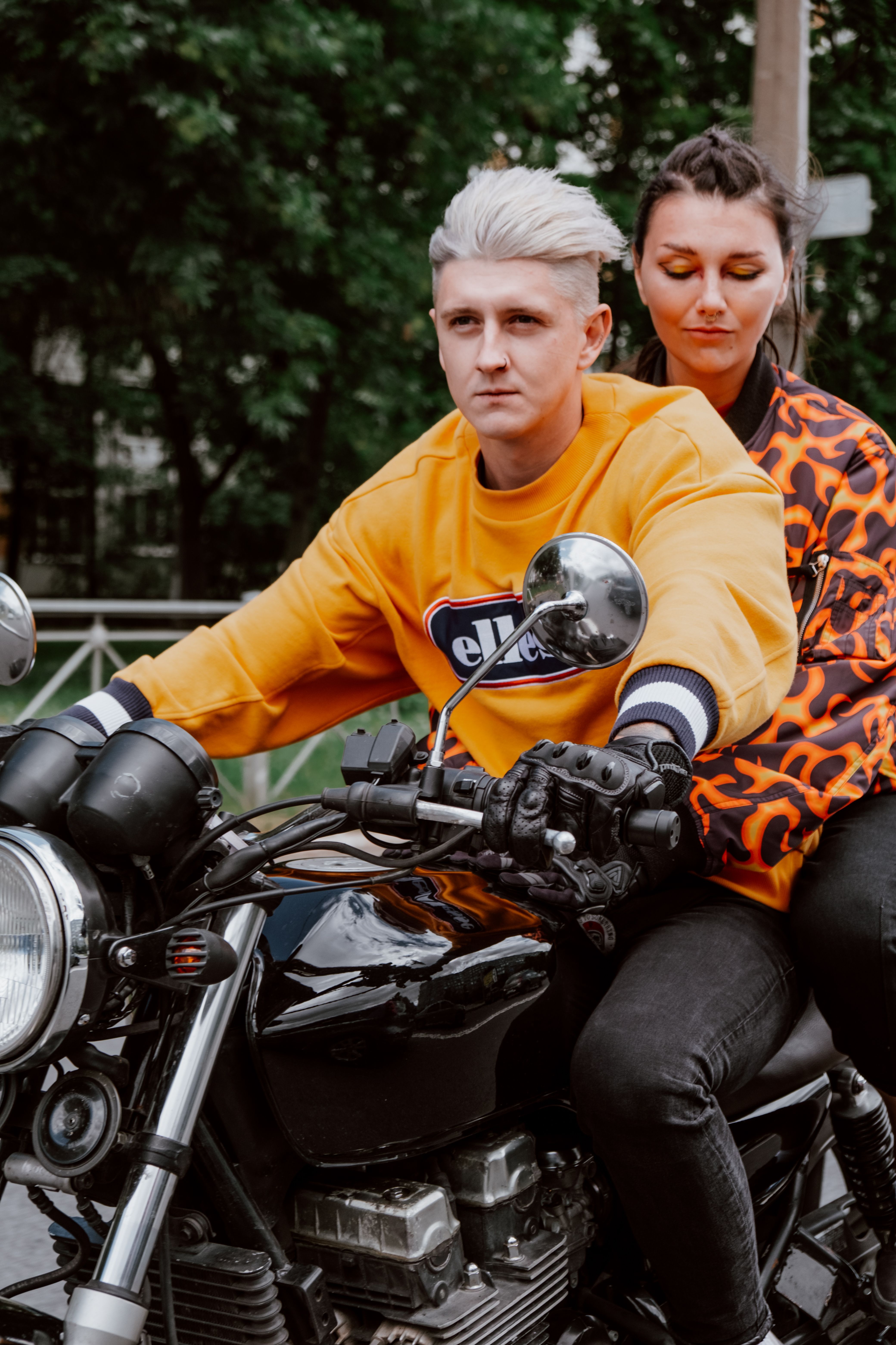 A stylish couple wearing vibrant jackets riding a classic motorcycle through an urban environment.