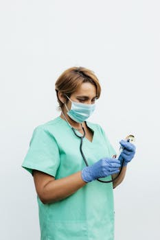 Healthcare professional in scrubs wearing mask and gloves, holding a stethoscope against a white background.