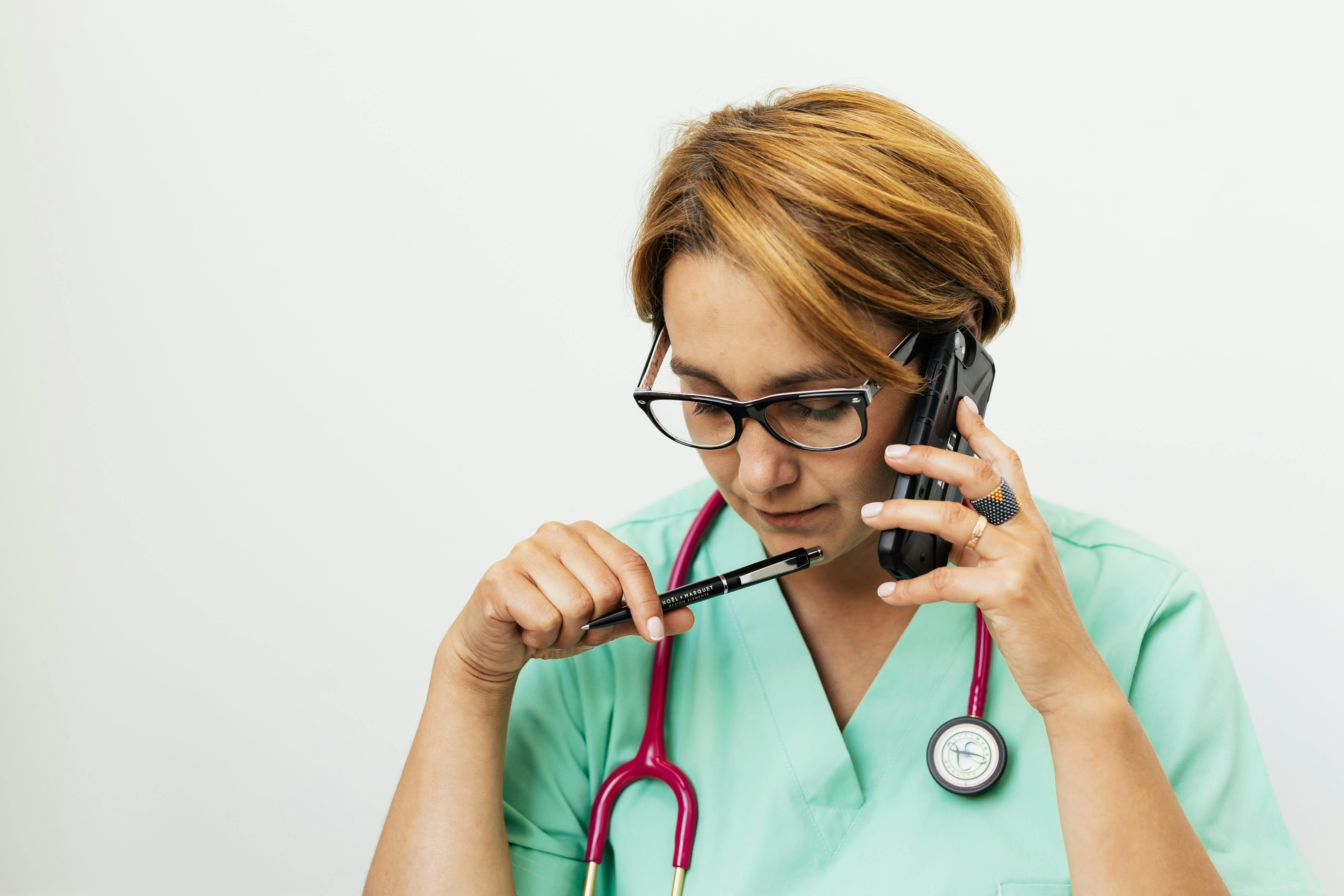 Healthcare professional in scrubs with stethoscope taking phone call while holding pen, depicting the importance of medical communication about health products.