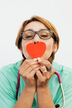 Female healthcare worker with eyeglasses holding a red paper heart, symbolizing care and compassion.