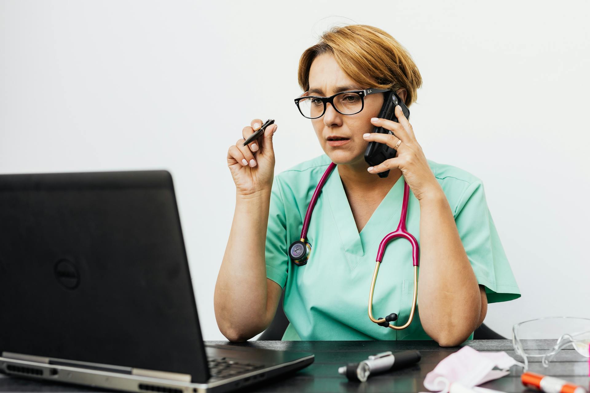 A woman in medical scrubs working with a laptop and phone, illustrating modern healthcare communication.