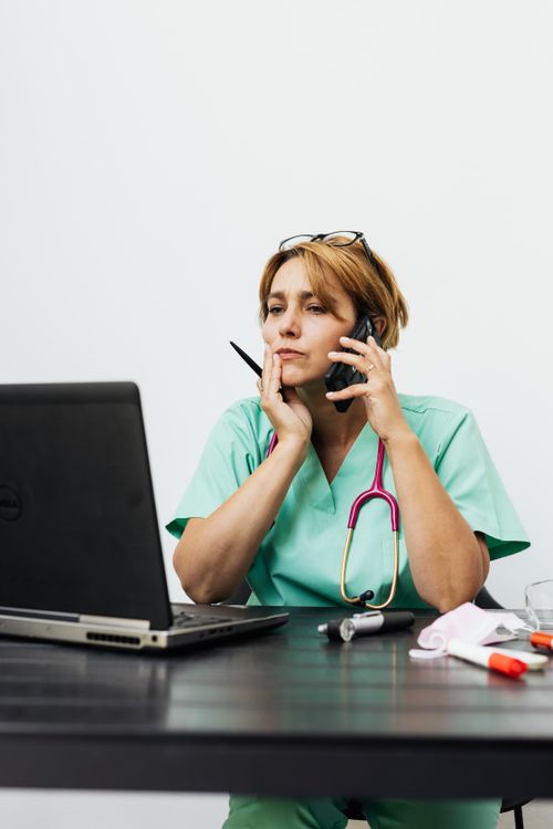 Free Doctor in green scrubs working remotely, using laptop and phone. Medical tools on table. Stock Photo