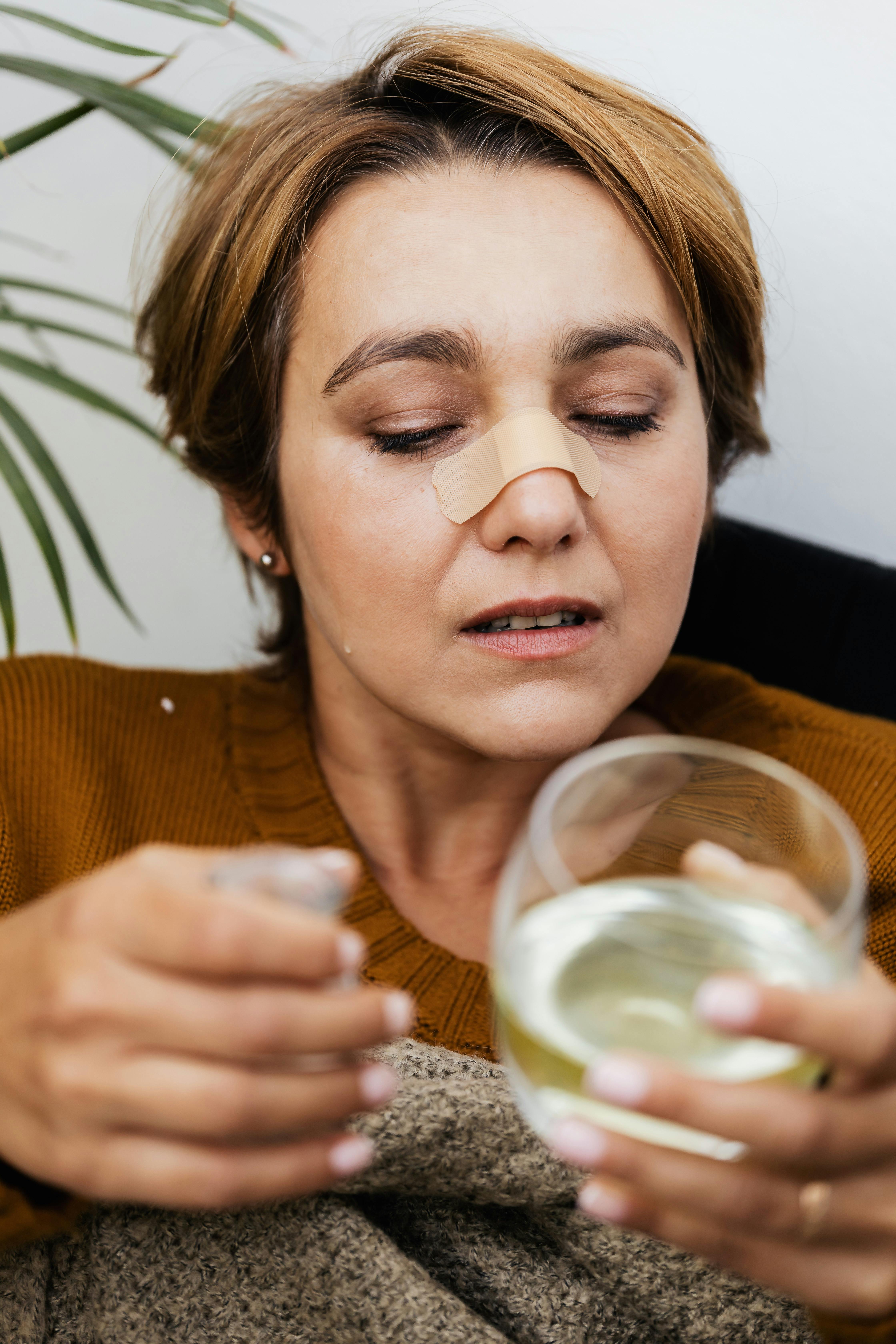 Adult woman with a bandage on her nose holding a glass of water, indoors.