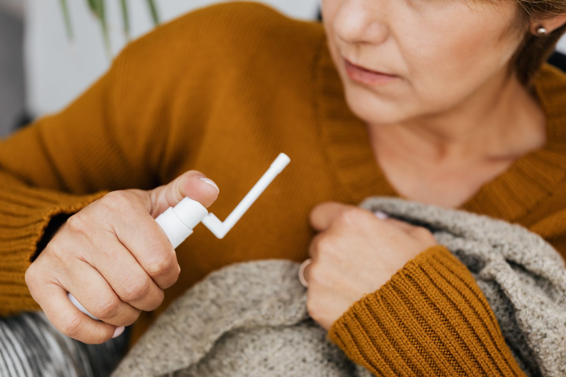Adult woman using a mouth spray for relief indoors, wrapped in a cozy sweater.