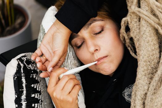 一名妇女裹着舒适的毯子躺在沙发上，用温度计测量体温