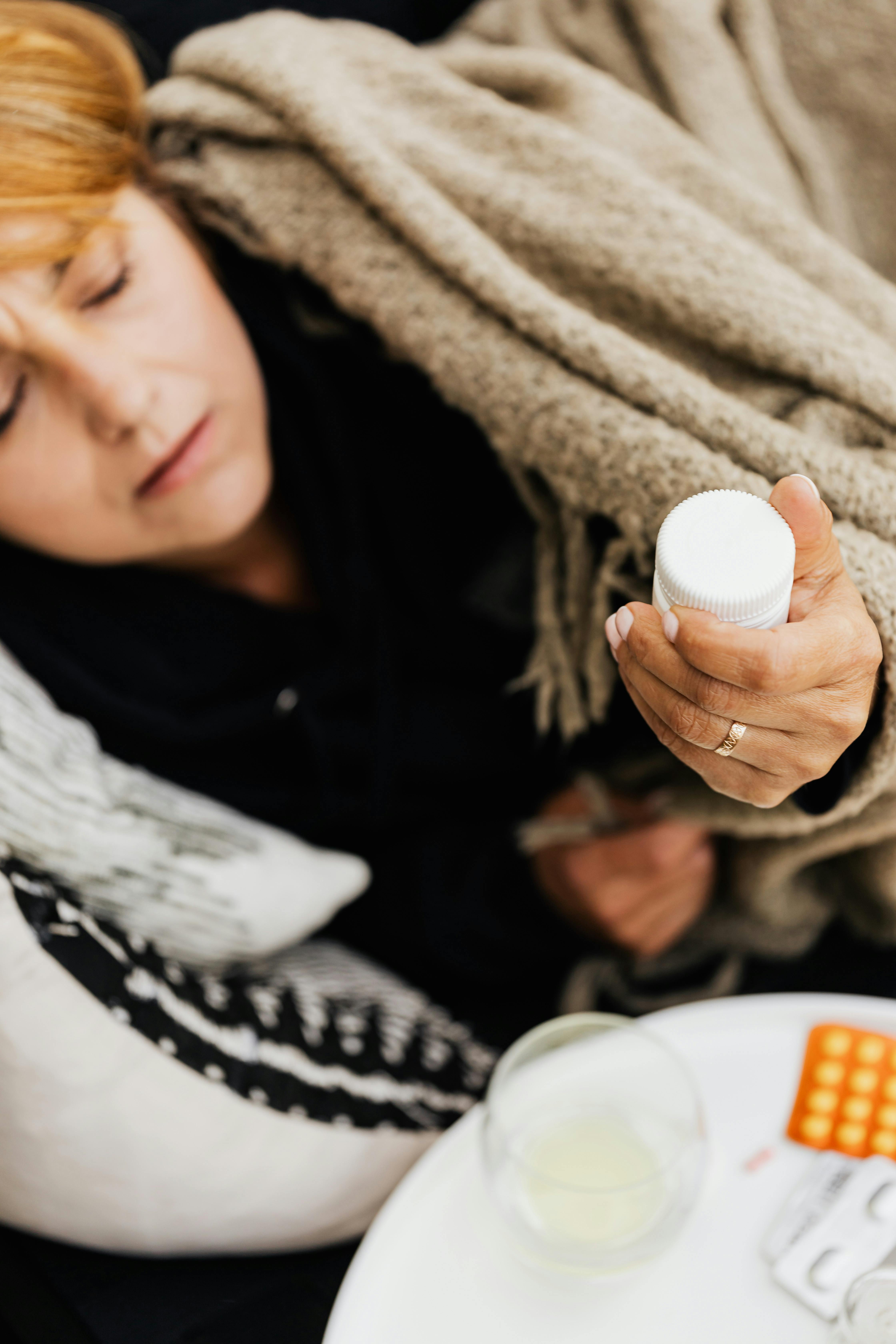 A woman wrapped in a blanket holding a medicine bottle, appearing ill at home.