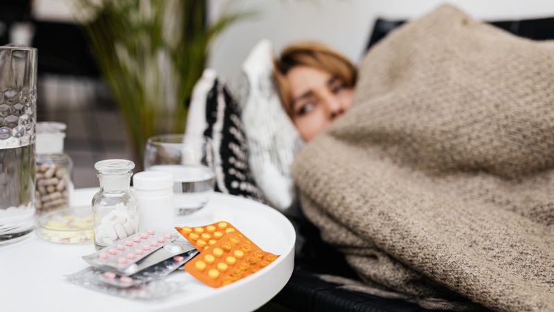 A woman rests under a blanket surrounded by medications on a table, depicting illness and recovery.