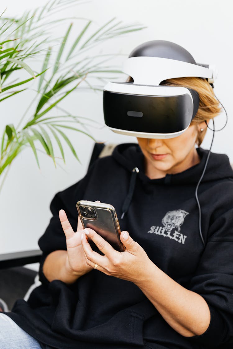 
A Woman In A Black Hoodie Wearing A VR Headset While Using Her Smartphone