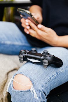 Person relaxing with a smartphone and video game controller on denim pants.