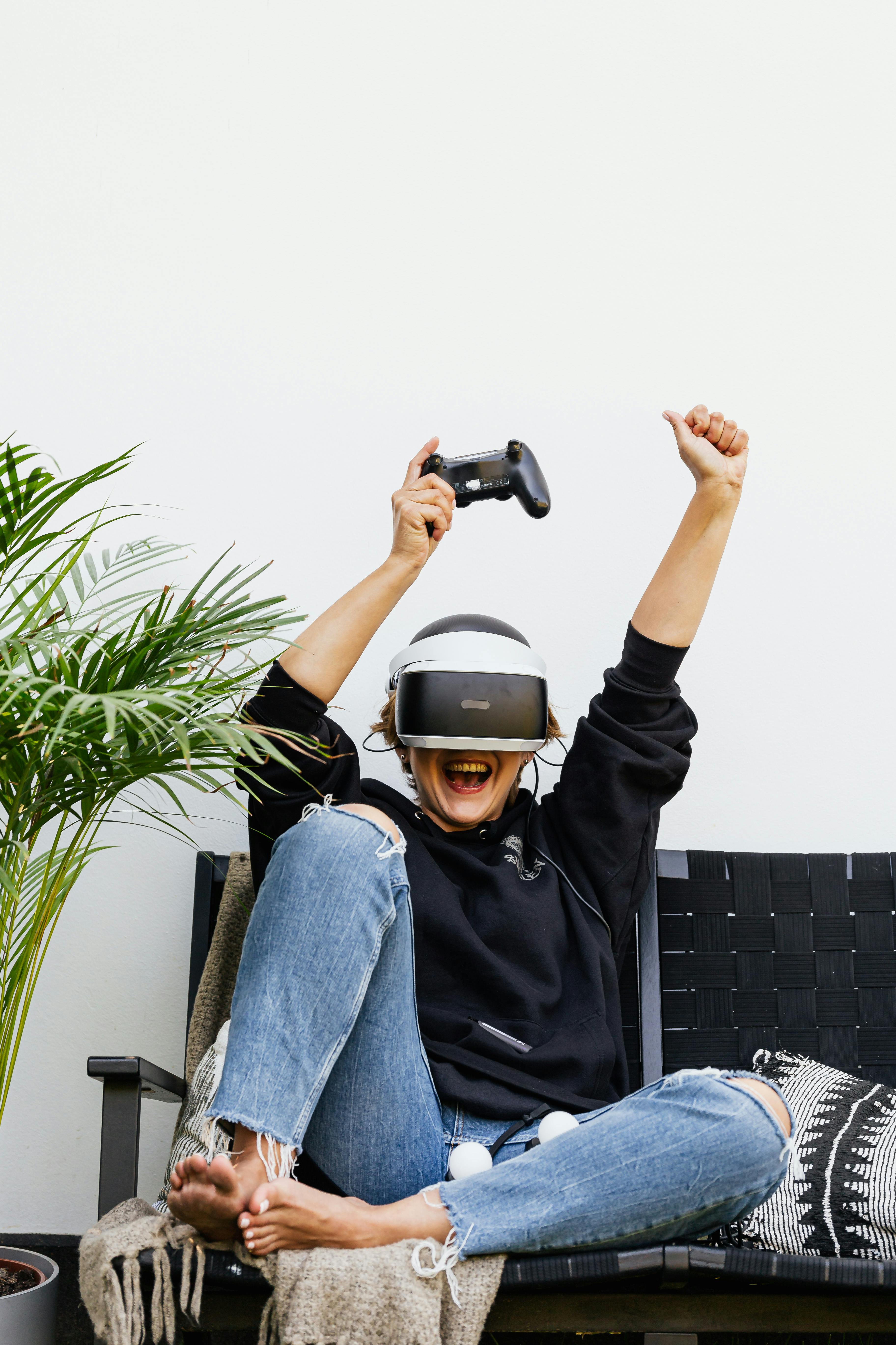 Woman in black hoodie celebrating while playing with VR headset and controller indoors.