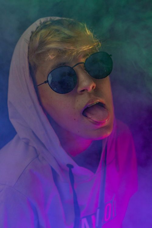 Free Confident female wearing hood and trendy sunglasses showing grimace with tongue out in neon light covered with smoke Stock Photo