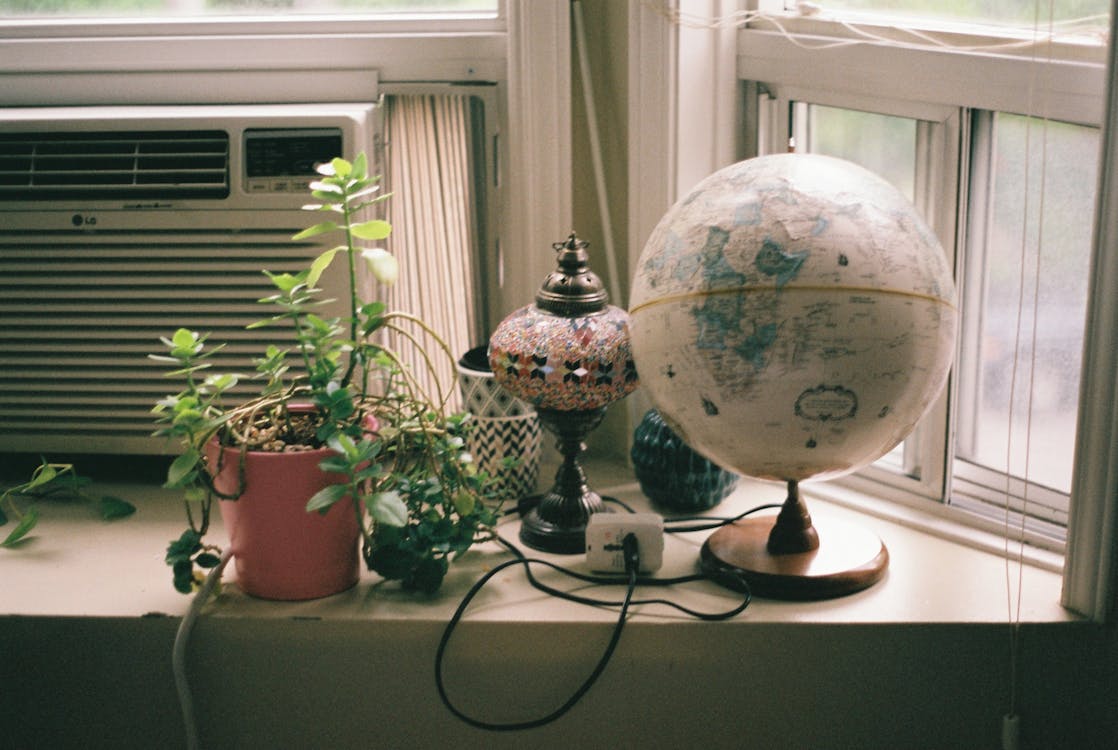 Free Wdecorativehite windowsill with potted plant and globe and decorative lamp placed near small window with air conditioner in light room at daytime inside Stock Photo