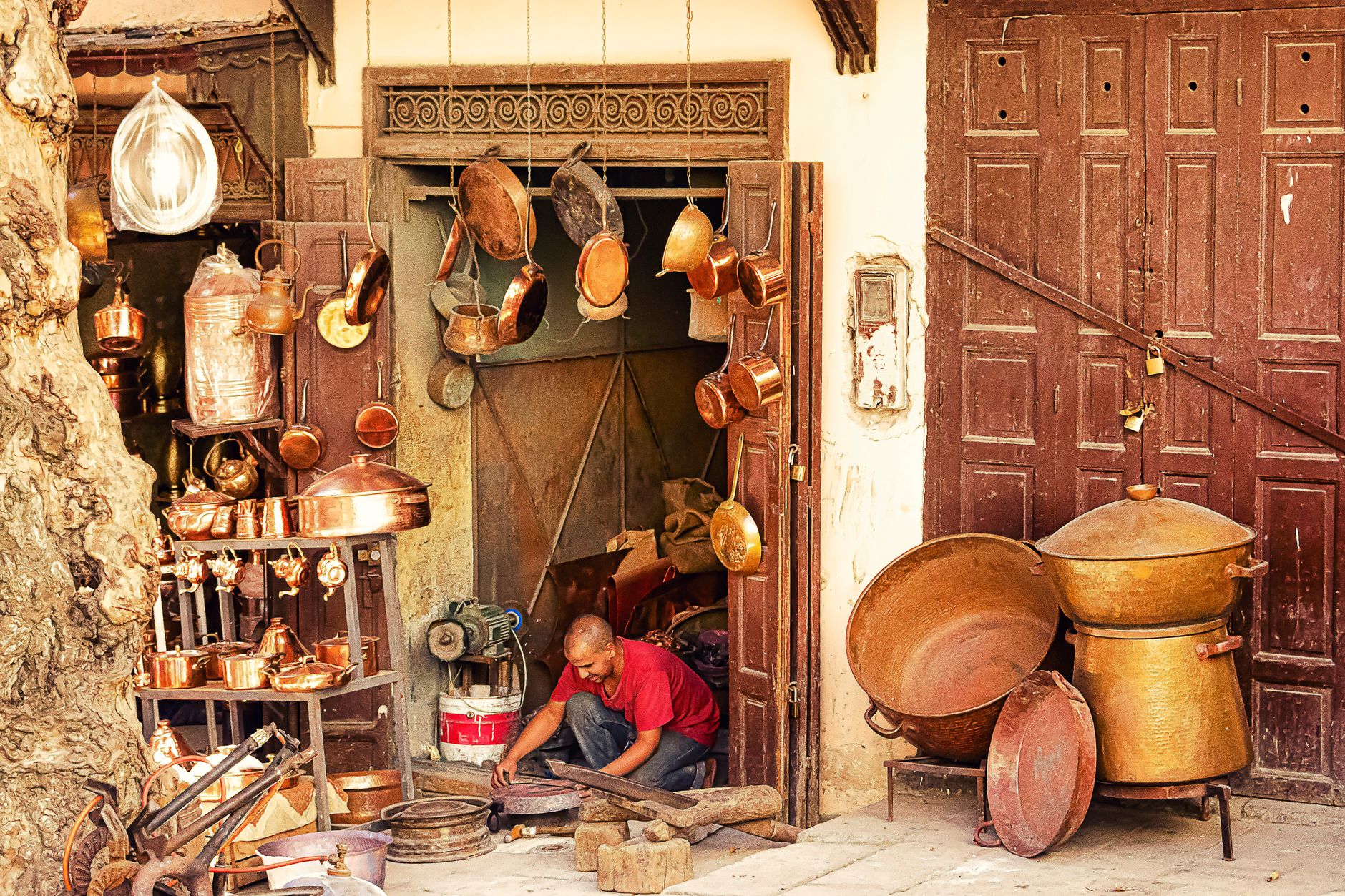 Coppersmith working on copperware outside a rustic workshop filled with pots and pans.