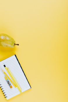 Flat lay of pear, notebook, and pencils on a bright yellow background.