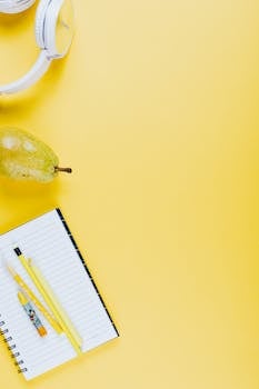 Top-view of a workspace featuring headphones, a pear, and a notepad on a yellow background.