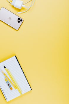 Flat lay of a smartphone, notebook, and pencils on a bright yellow background with ample copy space.