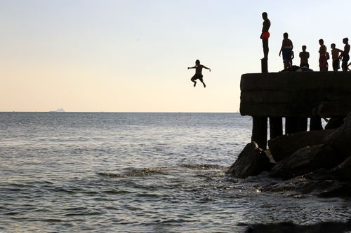 Free People silhouetted against the sky jump from a pier into the sea, capturing carefree summer fun. Stock Photo