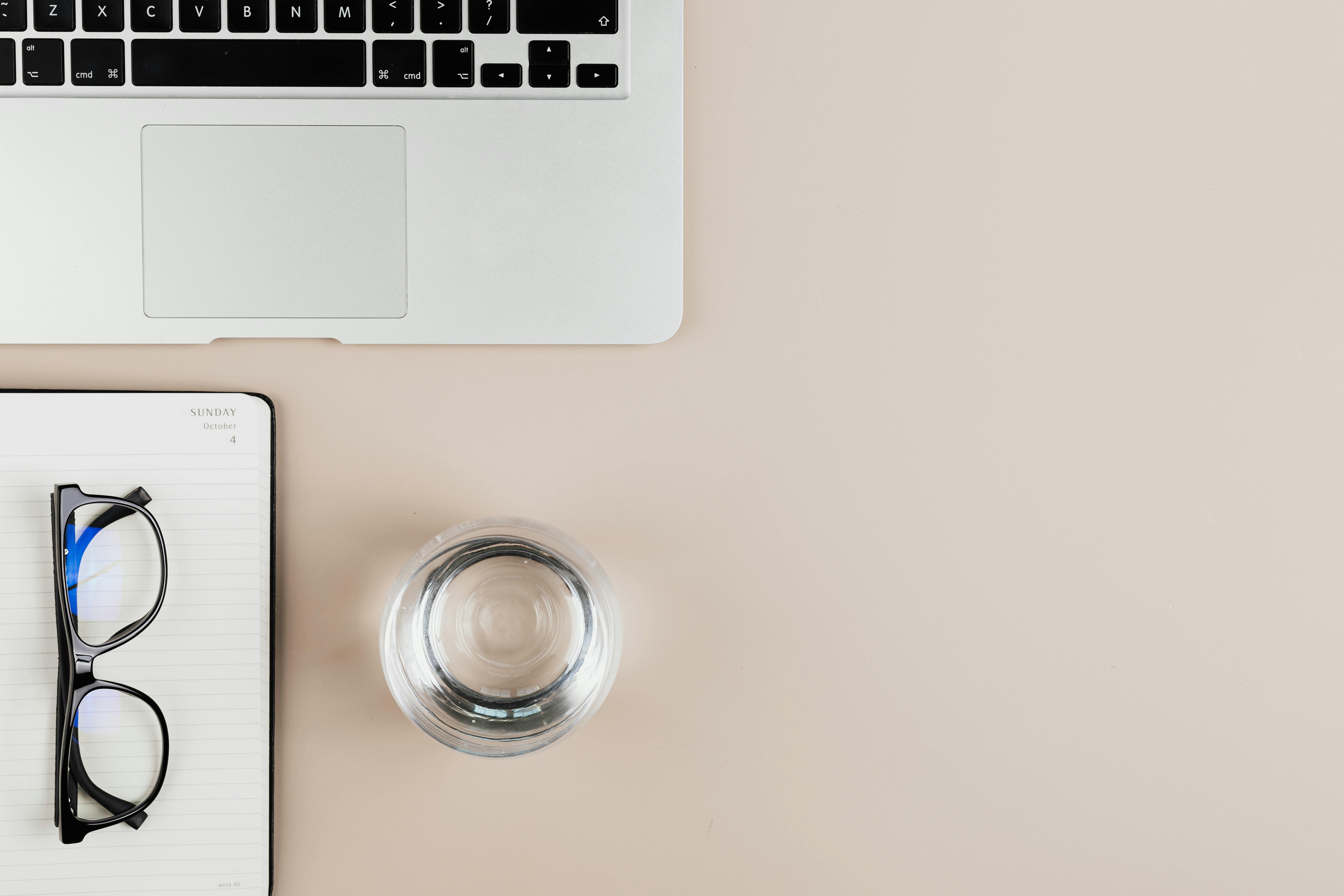 Overhead view of a clean workspace with a laptop, notebook, eyeglasses, and a glass of water.