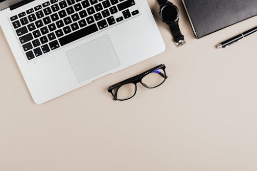 Modern workspace flat lay with laptop, notebook, watch, and glasses on a beige background.