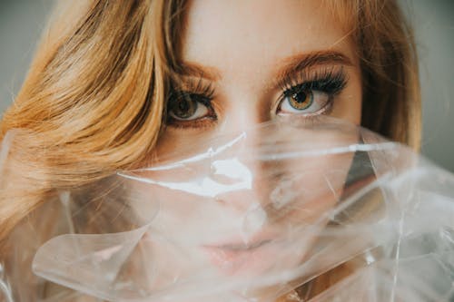 Serious female with blonde hair looking at camera while covering half face with transparent plastic package in light room inside