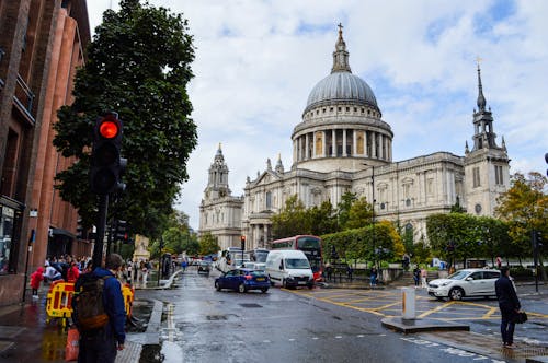 Exterior of aged stone cathedral on crowded wet street with hurrying people and cars on under blue cloudy sky