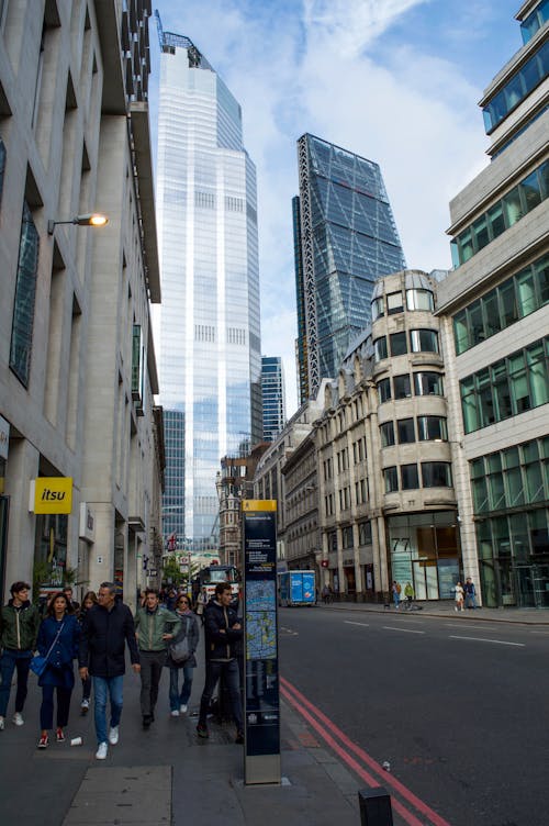 Low angle of unrecognizable pedestrians walking on city street near aged buildings and modern skyscrapers against cloudy blue sky