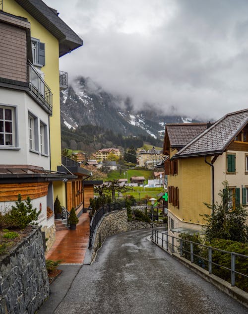 Free Picturesque scenery of narrow road amidst small village houses located in mountainous valley against foggy cloudy sky in Switzerland Stock Photo