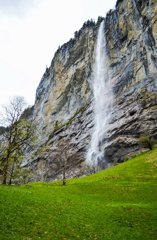 From below of picturesque Staubbach Fall waterfall flowing through rough rocky cliff and splashing near grassy hill slope against cloudy sky in Switzerland