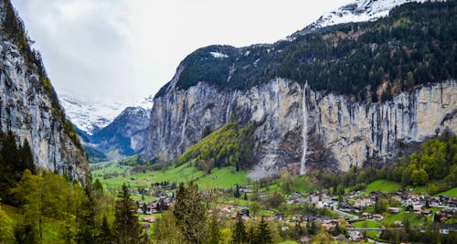 Free Magnificent alpine landscape of small Lauterbrunnen village located on grassy terrain between snowy rocky mountains and waterfall against cloudy sky in Switzerland Stock Photo
