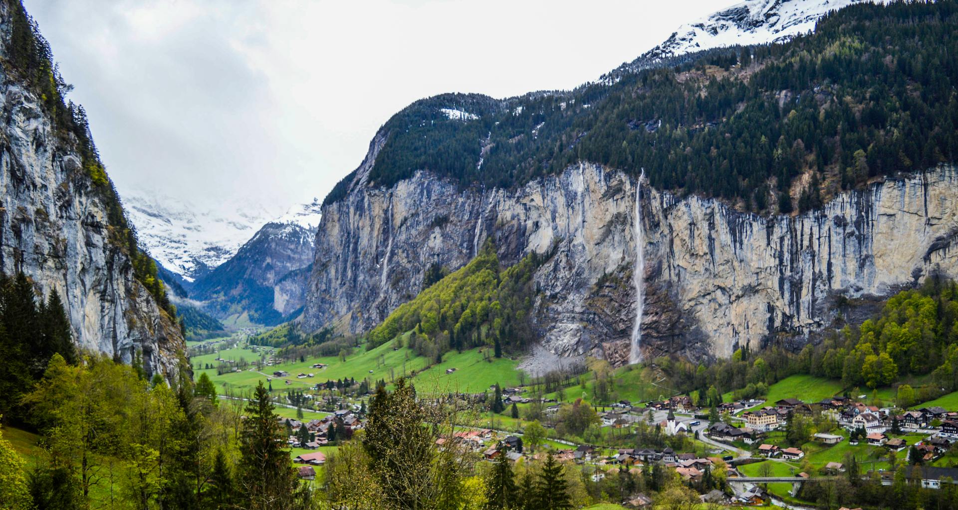 Magnificent alpine landscape of small Lauterbrunnen village located on grassy terrain between snowy rocky mountains and waterfall against cloudy sky in Switzerland