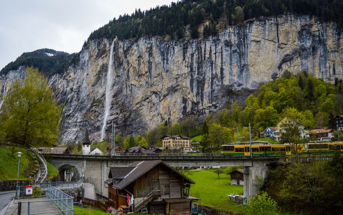 A yellow and green train crossing a bridge in the valley under the cliff, with the thin Staubbach falling behind it, chalets and a white church tower nearby, under an overcast sky