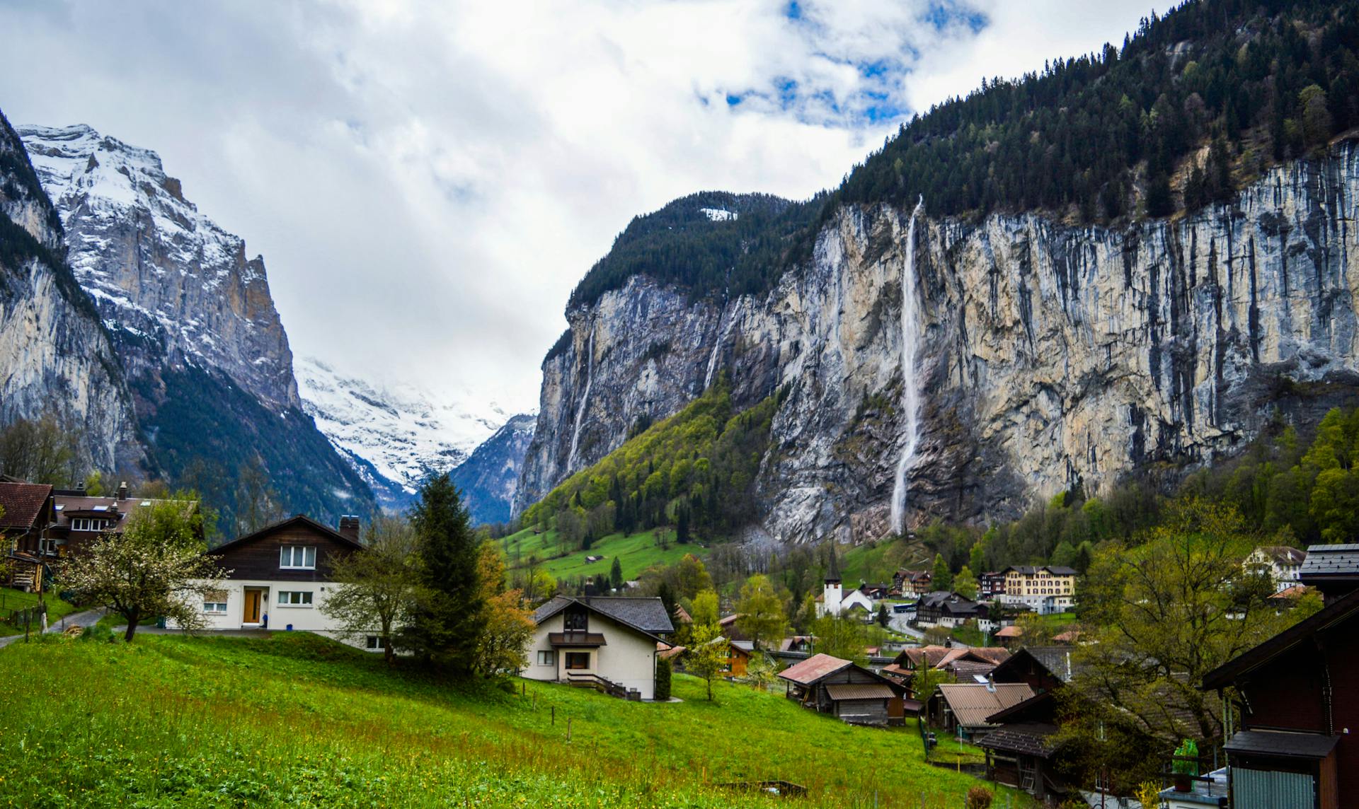 Traditional village houses located on green grassy terrain between huge rocky mountains with snow and trees on peaks in Lauterbrunnen