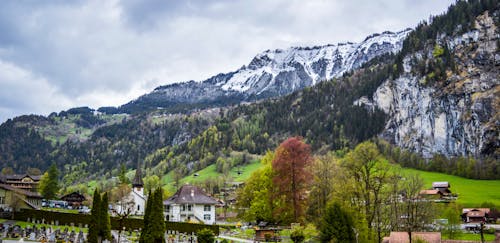 Free Breathtaking landscape of mountain ridge with lush green trees on slope and snow on peak surrounding small village in Switzerland against cloudy sky Stock Photo
