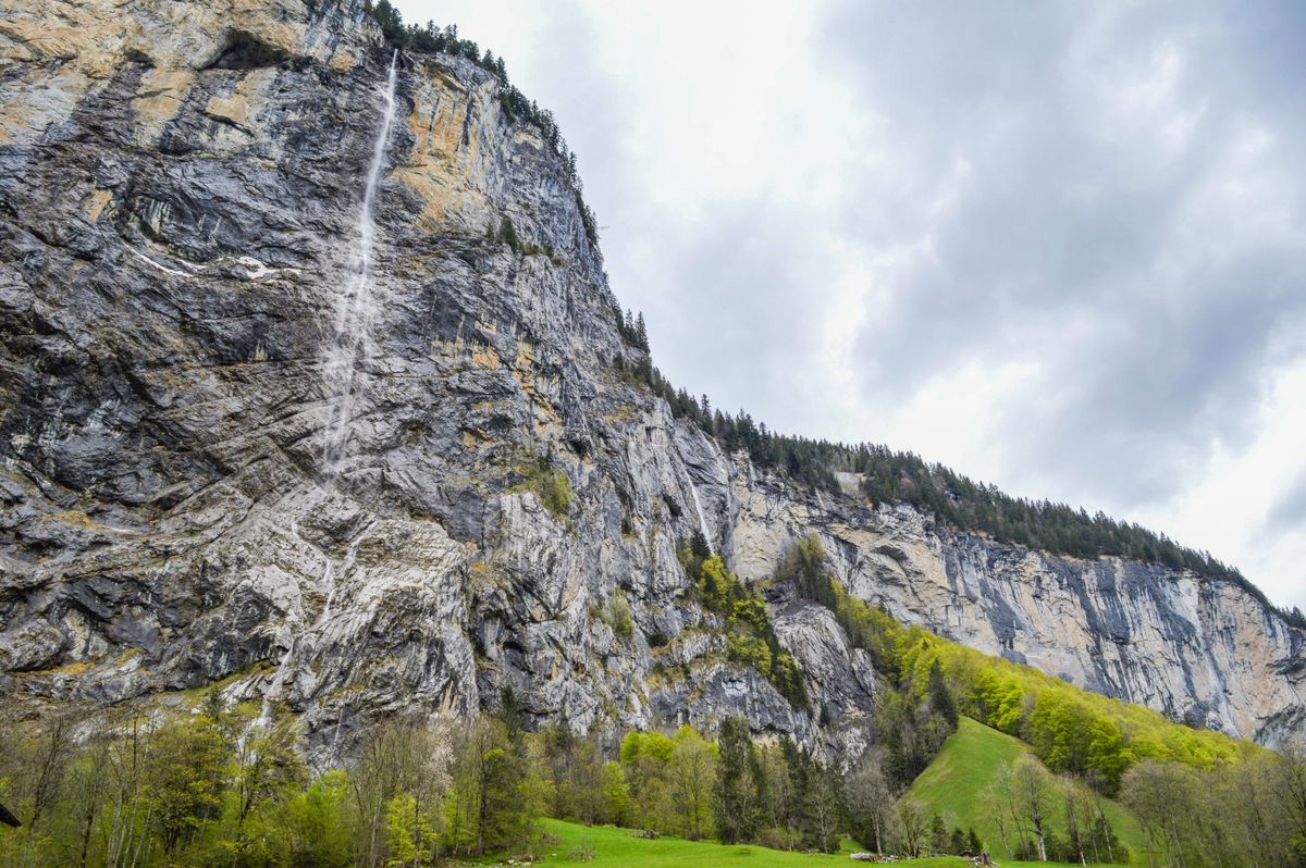 A long thin white ribbon of falling water down a huge grey rock wall, seen from a green meadow below, with conifers at the edge of the slope and a cloudy sky