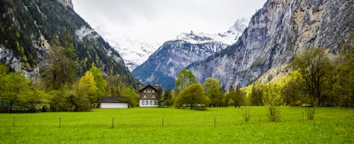 Free Amazing scenery of small residential house located on well groomed grassy meadow amidst magnificent massive mountains with snowy peaks in Switzerland Stock Photo