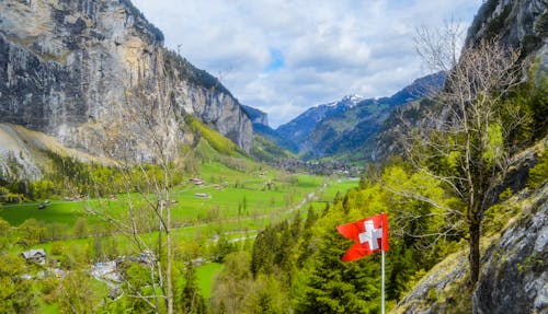 From above of small settlement located on green hill amidst highland with rocky cliff with flag of Switzerland in summer