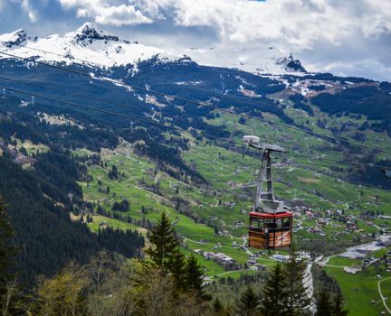 From above of cable car with little settlement below located oh grassy ground near mountains with snowy peaks in highlands