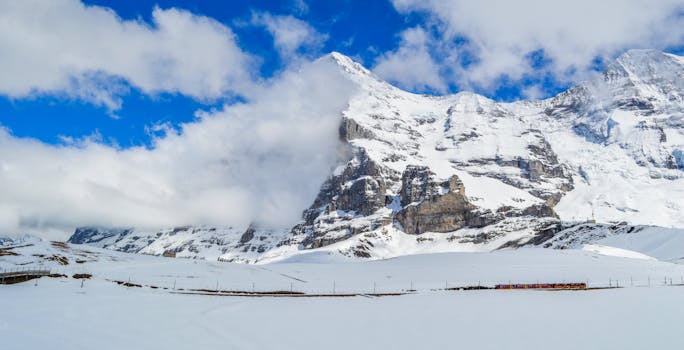 From below picturesque view of high snowy ridge near railway with train under bright sky with clouds in wintertime