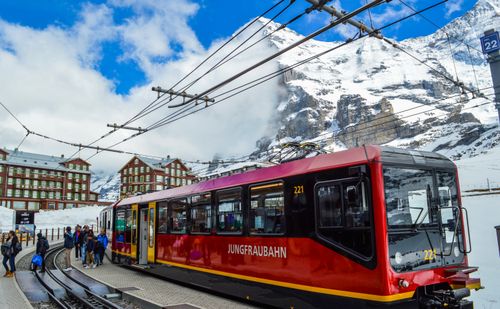 Free From below of contemporary train on railway near group of anonymous travelers on platform behind snowy mountain and buildings in winter Stock Photo