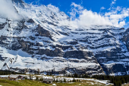 Spectacular alpine landscape of small settlement located on grassy terrain near massive rocky mountain covered with snow in Switzerland