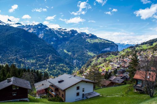 Free Picturesque scenery of village placed on grassy hill near mountains covered by green forest under blue cloudy sky Stock Photo