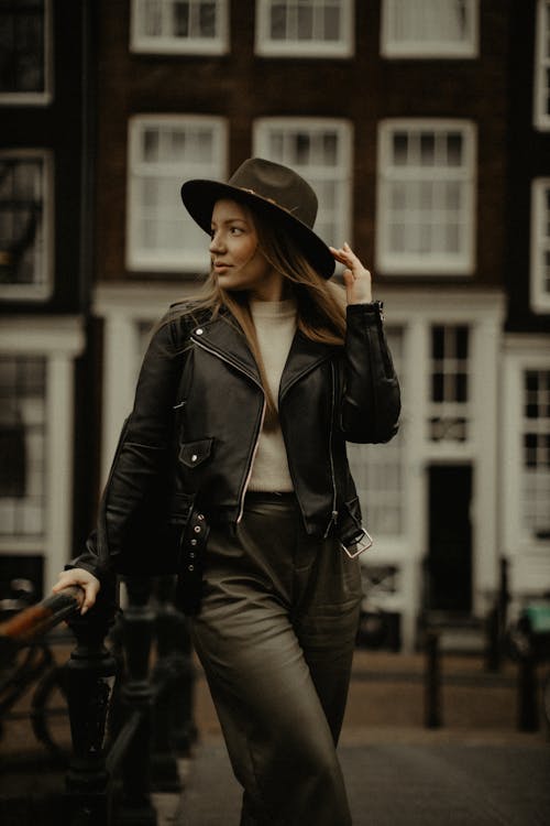 Free Beautiful female with long hair looking away while standing with hand on hat near metal railing and facade of building on blurred background Stock Photo