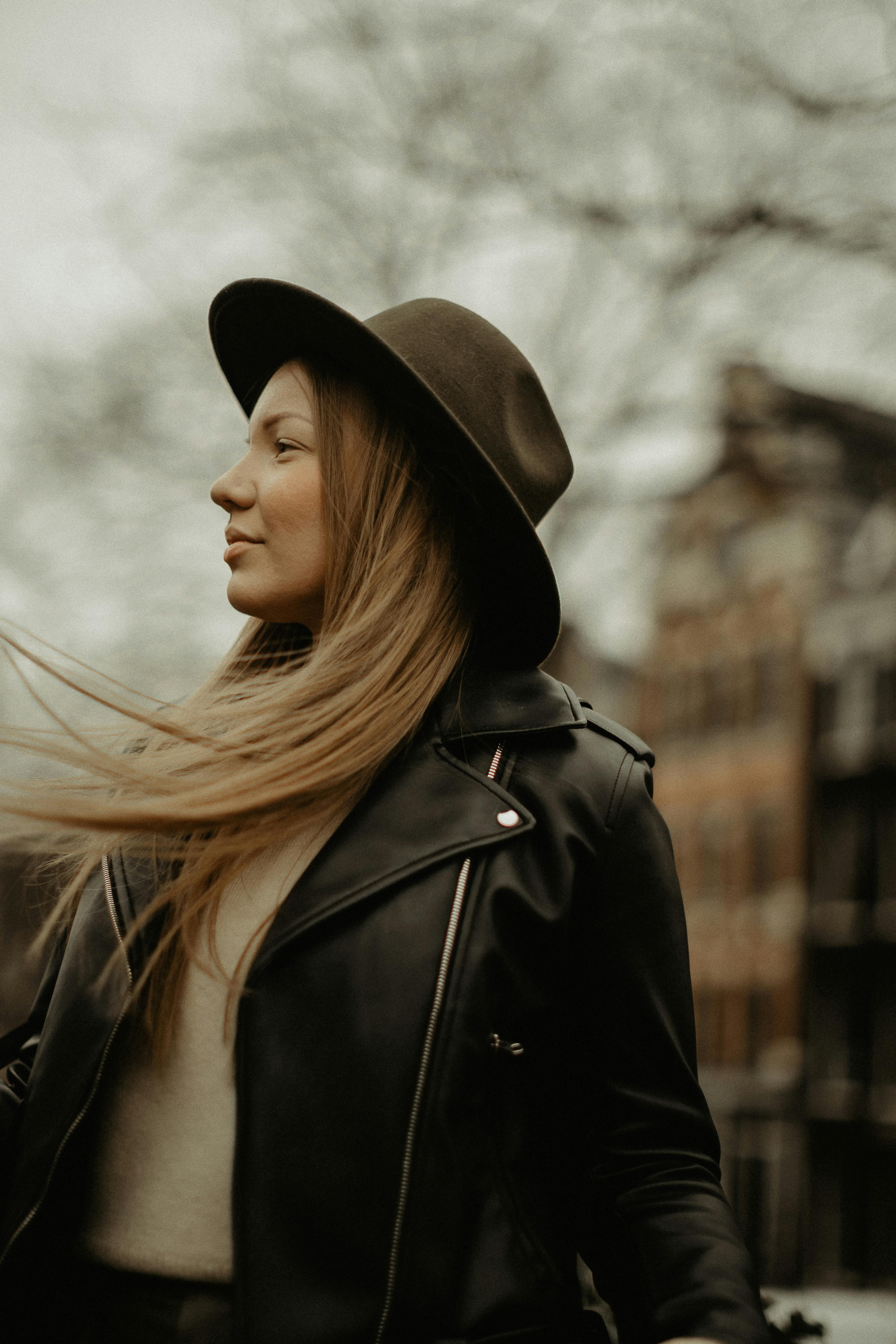 Side view of attractive female in hat and leather jacket standing on street with flowing hair and building on blurred background