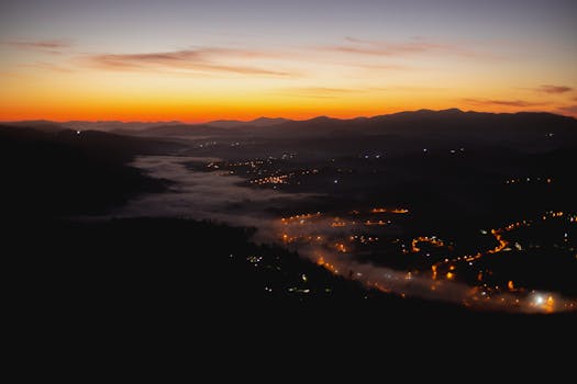A stunning aerial view of a foggy mountain village illuminated by city lights at sunset.