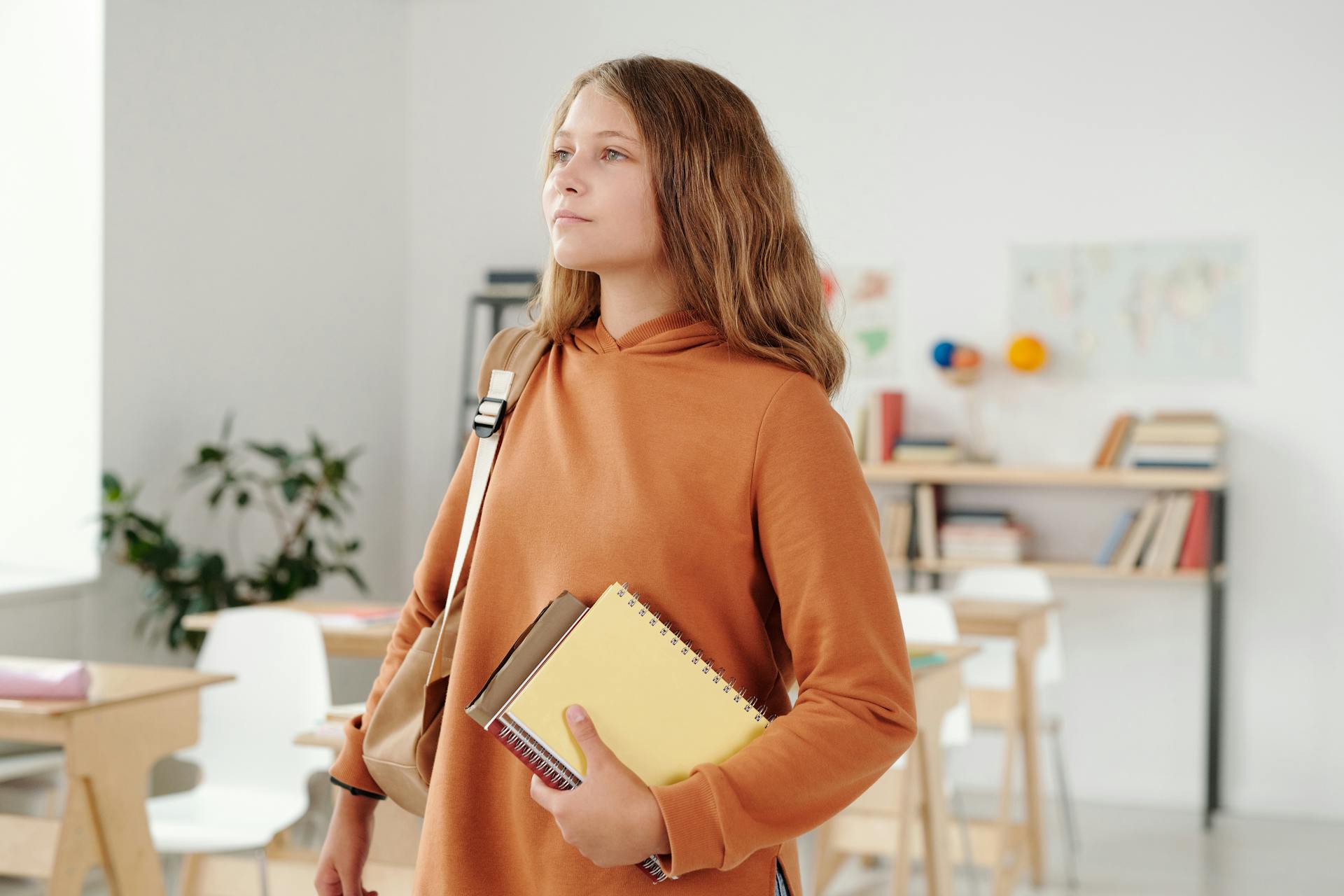 A student in a classroom holding books and a backpack, looking thoughtful.