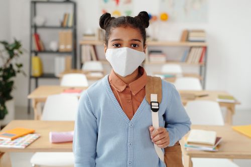 Free Girl with backpack and face mask standing in a classroom, symbolizing back-to-school safety. Stock Photo