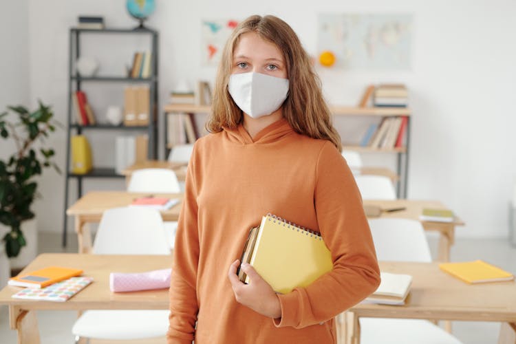 Kid Wearing Brown Sweater And Facemask Standing Inside A Classroom