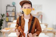 金发男孩背着书包，戴着帽子和面具，准备去室内上学