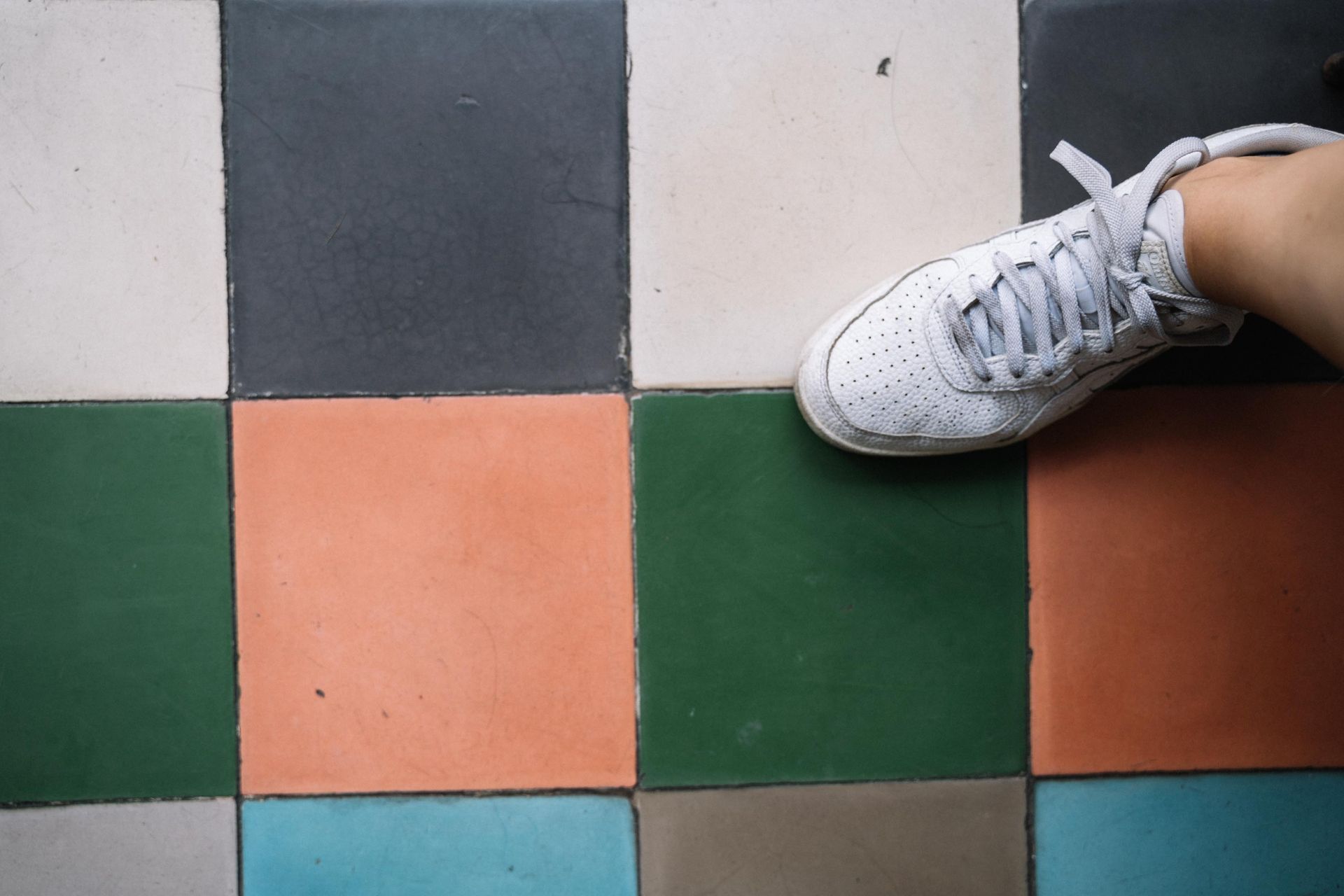 A white sneaker stepping on a colorful tiled floor featuring green, orange, and blue tiles.